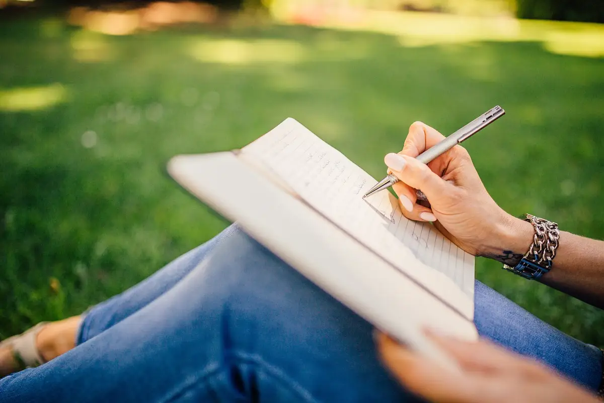 A person rests a notebook on their knees. They are wearing jeans in front of a green background. We can see their hands. They are writing in the notebook.