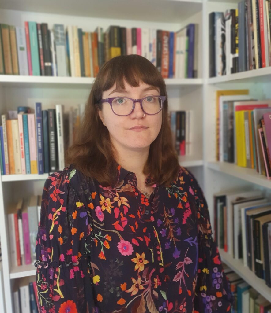 Flo is a white person with mid-length brown hair and glasses. They are wearing a black shirt patterned with colourful flowers, and standing in front of some white bookshelves filled with books. They are looking at the camera and slightly smiling.