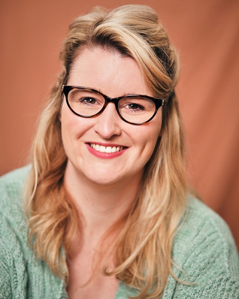 Jade has long blonde hair and is wearing glasses with a thick black frame. She is smiling at the camera, wearing a pale green top in front of an orange background.