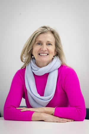 Lucy is a person with shoulder-length blonde hair. She is wearing a bright pink jumper and a pale blue scarf. She is looking at the camera and smiling.