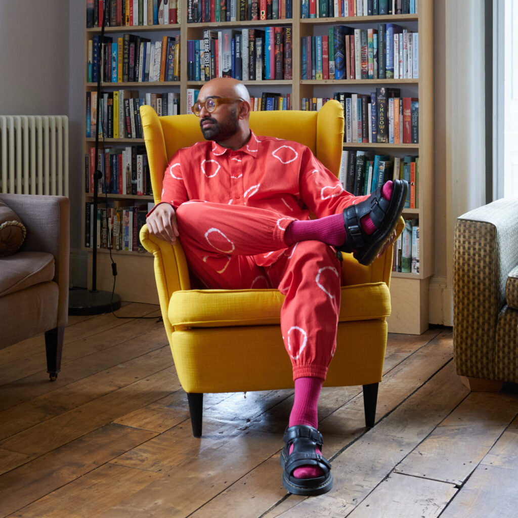 Karrish is wearing a suit of red, patterned fabric. He is sitting in a yellow armchair in front of a bookshelf. He is looking away from the camera, wearing glasses and a slight smile.