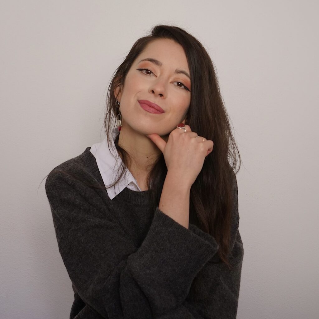 Nazaret has long brown hair and is wearing a black jumper over a pale collared shirt. She is sitting in front of a white background and smiling at the camera, with her chin resting on one hand.