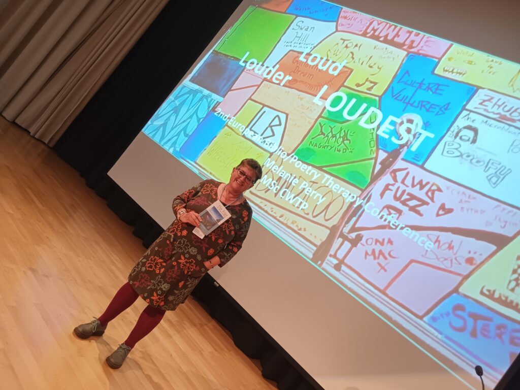 Mel Perry stands before a large slide projection for her presentation  "Loud, Louder, Loudest" at the 2nd European Biblio/Poetry Therapy Conference in Jyväskylä, Finland. The slide is colourful patchwork of image and text. Mel is wearing a patterned dress and smiling at the camera.