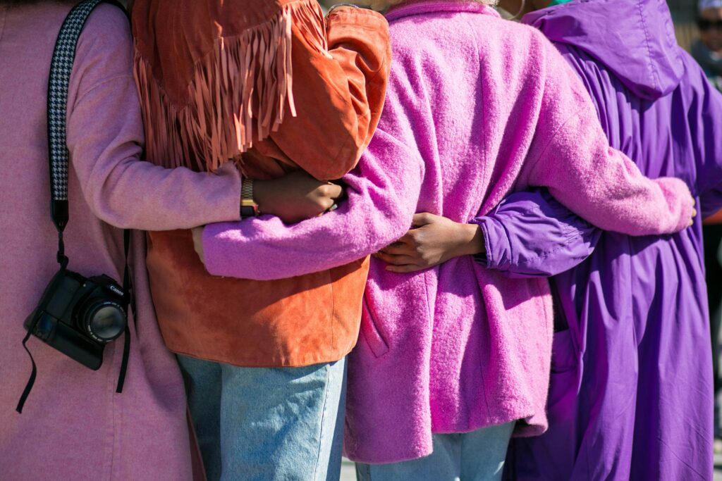 For women are seen from behind. They have their arms around each other in a line or circle. They are wearing colourful coats and jackets in pink, orange and purple.